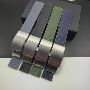 May include: A collection of watch straps in various colors, including gray, black, olive green, and navy blue. Each strap features a textured surface and a silver-colored metal clasp. The straps are arranged side by side, showcasing their different hues and designs.