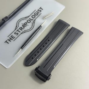 May include: A black rubber watch strap with a deployment clasp, a metal spring bar tool, and three silver spring bars. The items are arranged next to a translucent plastic pouch featuring the black text THE STRAPOLOGIST and a clock icon.