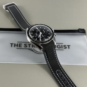 Könnte beinhalten: Eine Omega Speedmaster Chronographenuhr mit schwarzem Zifferblatt, drei Hilfszifferblättern und einem schwarzen, strukturierten Armband mit weißen Kontrastnähten. Die Uhr liegt auf einer durchscheinenden Kunststofftasche mit dem Aufdruck THE STRATEGIST.