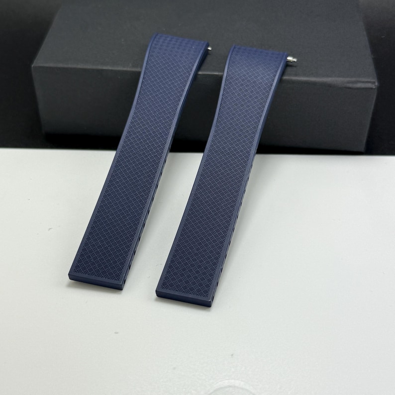 May include: Two navy blue watch straps with a textured, diamond-patterned surface. The straps are curved and have silver-colored pins at one end for attachment. The straps are lying on a white surface with a black box in the background.