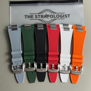 Puede incluir: Seis correas de reloj en gris, verde, negro, rojo, blanco y naranja, cada una con una superficie texturizada, una hebilla de metal plateado y un conector metálico. Las correas se muestran frente a una bolsa de plástico transparente con la etiqueta THE STRAPOLOGIST.