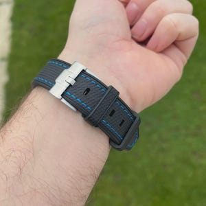 Puede incluir: Una correa de reloj negra con superficie texturizada y costuras en contraste de color azul brillante. La correa cuenta con un cierre desplegable de metal plateado y dos trabillas de goma negra a juego para asegurar la correa del reloj.