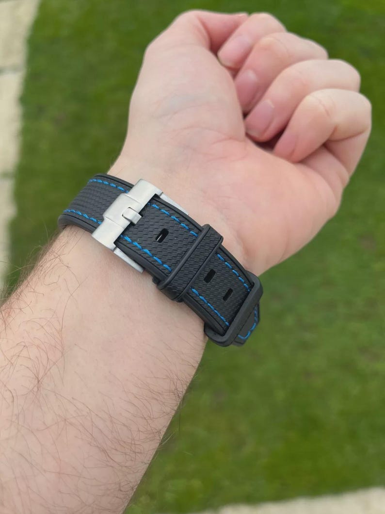 Puede incluir: Una correa de reloj negra con superficie texturizada y costuras en contraste de color azul brillante. La correa cuenta con un cierre desplegable de metal plateado y dos trabillas de goma negra a juego para asegurar la correa del reloj.
