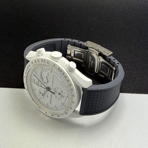 May include: A white wristwatch with a gray band. The watch face displays multiple dials and the word "Speedmaster". The band is gray with a textured pattern. The clasp is stainless steel.