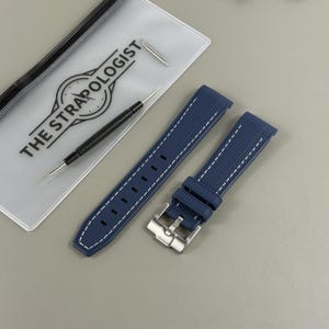 Puede incluir: Correa de reloj azul marino con costuras blancas y hebilla plateada. La correa está junto a una bolsa transparente con el texto "THE STRAPOLOGIST" y una herramienta de reloj.