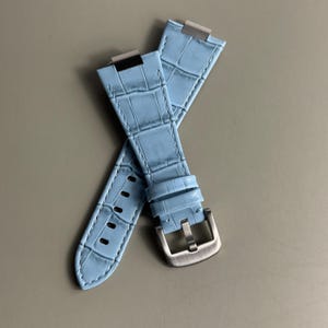 Puede incluir: Una correa de reloj azul claro con un patrón texturizado de relieve de cocodrilo. La correa incluye una hebilla de metal plateado cepillado y herrajes de fijación metálicos integrados en los extremos, con costuras tonales visibles.