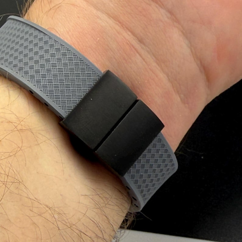 May include: Close-up of a gray watch band with a woven pattern and a black clasp. The band is made of a flexible material and is secured with a rectangular black clasp. The watch band is on a wrist.