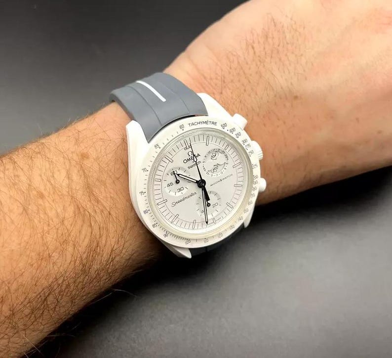 Może przedstawiać: Biały zegarek naręczny chronograf z szarym paskiem z gumy. Tarcza zegarka posiada skalę tachymetru, okienko daty oraz nadrukowane słowa "Speedmaster" i "Omega".