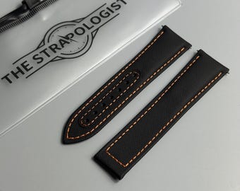 May include: A black watch strap with orange contrast stitching, featuring a textured surface and multiple adjustment holes. The strap is positioned next to a clear plastic packaging sleeve printed with the text THE STRAPOLOGIST.