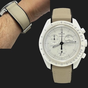 May include: A white Omega Speedmaster MoonSwatch watch with a beige canvas strap. The watch face features a tachymeter scale, chronograph subdials, and the text "OMEGA swatch" and "MOON SWATCH".