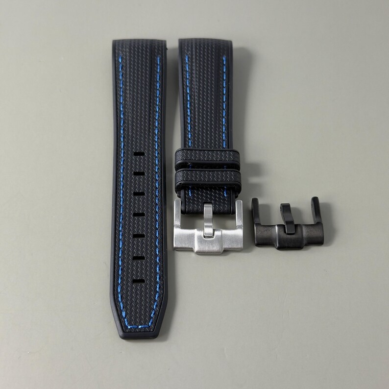 Puede incluir: Una correa de reloj negra con un patr&oacute;n de fibra de carbono texturizado y costuras azules. La correa incluye dos trabillas, una hebilla de metal plateado y un componente de hebilla de metal negro adicional para uso intercambiable.