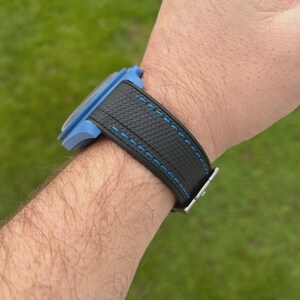 Puede incluir: Un reloj de pulsera con caja azul claro y correa negra texturizada con costuras en azul brillante. La correa presenta un patr&oacute;n de rayado cruzado distintivo y se asegura con una hebilla de metal plateado que contrasta con la caja azul.