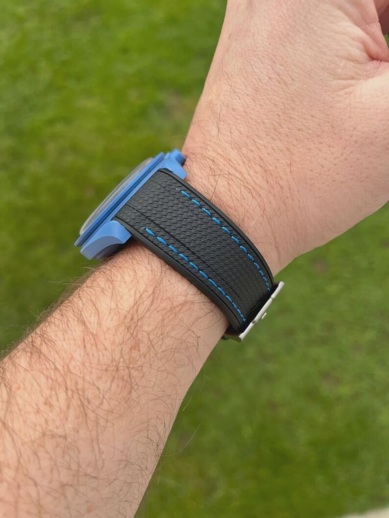 Puede incluir: Un reloj de pulsera con caja azul claro y correa negra texturizada con costuras en azul brillante. La correa presenta un patr&oacute;n de rayado cruzado distintivo y se asegura con una hebilla de metal plateado que contrasta con la caja azul.