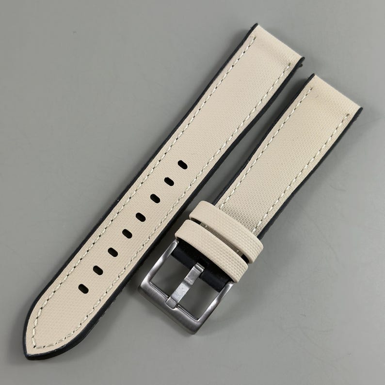 May include: Cream-colored watch strap with black backing and white stitching. The strap features a stainless steel buckle and multiple adjustment holes. The watch band is made of a textured canvas material.