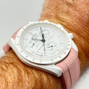 May include: White Omega Speedmaster MoonSwatch watch with a white face, black hands, and a pink strap. The watch has the Omega and MoonSwatch logos, and the word "Speedmaster" is printed on the face.