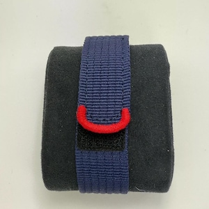 Puede incluir: Una correa de reloj de nailon tejido azul marino con un llamativo detalle semicircular cosido en rojo cerca del extremo. La correa est&aacute; envuelta alrededor de un coj&iacute;n de exhibici&oacute;n rectangular gris oscuro con textura aterciopelada.