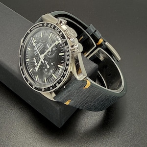May include: A silver-toned wristwatch with a black face and a black leather band. The watch face features white numerals and hands, with the brand name "Omega" visible. The band has yellow accents and is secured with a silver buckle.