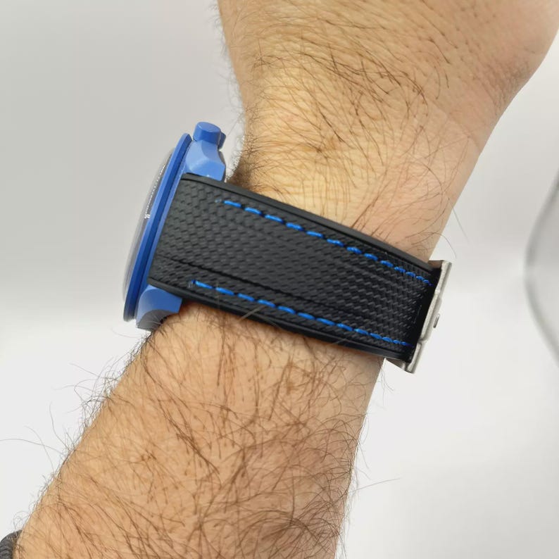 Puede incluir: Un reloj de pulsera con una caja azul brillante y una correa negra texturizada con costuras en azul vibrante. El reloj se muestra de perfil, destacando la corona azul y el patr&oacute;n detallado del material de la correa sobre un fondo liso.