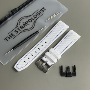 May include: A white textured watch strap with blue stitching, a silver-toned metal buckle, and a black metal buckle. Included are a black spring bar tool, two spring bars, and a translucent carrying case labelled THE STRAPOLOGIST.