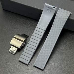 May include: A set of gray watch straps and a silver-tone clasp. One strap has a textured pattern, the other has a ribbed design. The text "XO-20" is visible on one strap. The watch accessories are displayed on a dark surface.