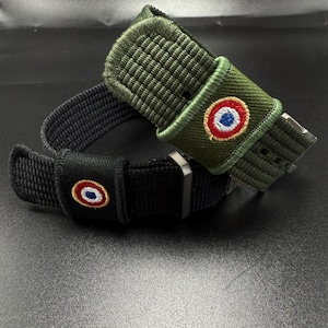 Puede incluir: Dos correas de reloj, una en verde oliva y otra en negro, ambas con textura de tela tejida y hebilla de metal. Cada correa incluye un peque&ntilde;o parche bordado circular que muestra un dise&ntilde;o de c&iacute;rculos conc&eacute;ntricos en rojo, blanco y azul.