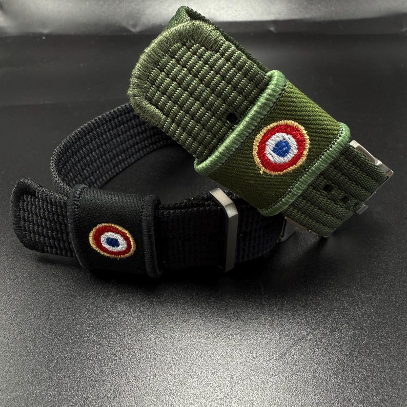Puede incluir: Dos correas de reloj, una en verde oliva y otra en negro, ambas con textura de tela tejida y hebilla de metal. Cada correa incluye un peque&ntilde;o parche bordado circular que muestra un dise&ntilde;o de c&iacute;rculos conc&eacute;ntricos en rojo, blanco y azul.