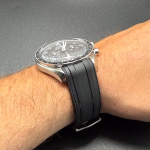 Puede incluir: Un reloj de pulsera de acero inoxidable con esfera negra y correa de caucho negra. El reloj presenta m&uacute;ltiples subesferas y un bisel con escala taquim&eacute;trica. El reloj se sujeta a la mu&ntilde;eca con una correa negra.