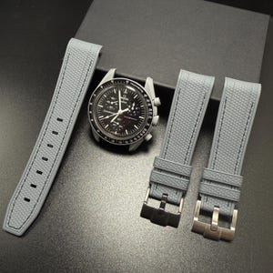 May include: A black-faced wristwatch with a silver bezel and grey straps. The watch has multiple dials and a tachymeter scale. Two grey watch straps with silver buckles are next to the watch, and one strap is to the left.