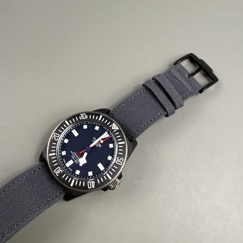 Puede incluir: Un reloj Tudor Pelagos Alinghi Red Bull Racing con esfera azul oscuro, marcadores de hora blancos y segundero rojo. El reloj cuenta con un bisel giratorio negro con n&uacute;meros blancos y una correa de tela gris con cierre de hebilla negro.