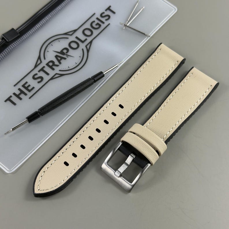 May include: A pair of cream-colored watch straps with black edges and white stitching. A silver-colored buckle is attached to one strap. The image also includes a watch tool and the text "THE STRAPOLOGIST".