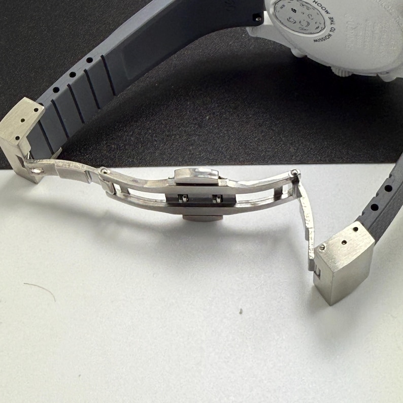 May include: Close-up of a luxury watch clasp and strap. The watch strap is dark gray, and the clasp is silver. The watch face is white with black text. The watch is on a black and white surface.
