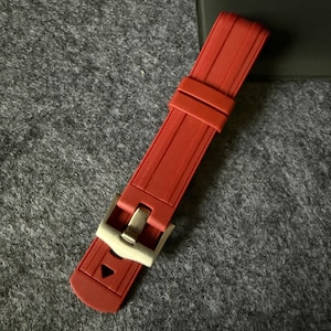 Puede incluir: Una correa de reloj roja con una hebilla plateada. La correa est&aacute; hecha de un material flexible y tiene una superficie texturizada. La hebilla es rectangular y tiene un dise&ntilde;o sencillo.