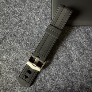 Puede incluir: Una correa de reloj negra con una hebilla plateada. La correa est&aacute; hecha de un material texturizado y acanalado. La hebilla tiene forma rectangular con un cierre. La correa del reloj est&aacute; sobre una superficie gris.