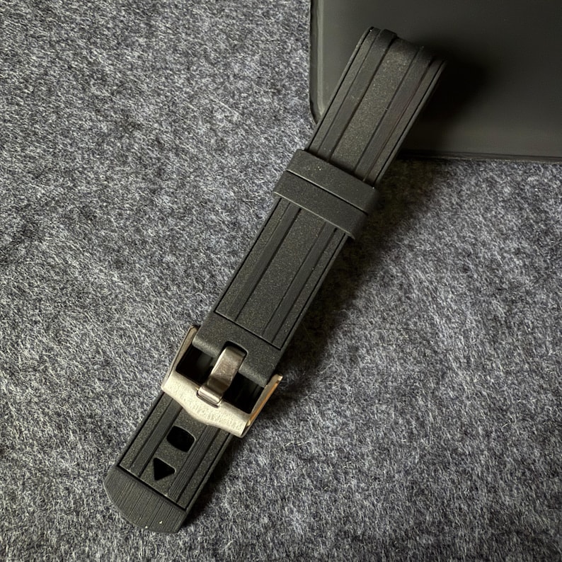 Puede incluir: Una correa de reloj negra con una hebilla plateada. La correa est&aacute; hecha de un material texturizado y acanalado. La hebilla tiene forma rectangular con un cierre. La correa del reloj est&aacute; sobre una superficie gris.