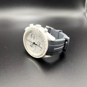 Może przedstawiać: Biały zegarek naręczny z chronografem i czarnym paskiem z gumy. Tarcza zegarka ma białe tło z czarnymi oznaczeniami i wskaz&oacute;wkami. Zegarek ma skalę tachymetru wok&oacute;ł zewnętrznej krawędzi tarczy.