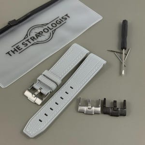 May include: Light gray watch strap with white stitching, a silver buckle, and a black and silver clasp. The strap has a textured pattern. The text "THE STRAPOLOGIST" is visible on a clear pouch. A watch tool is also included.