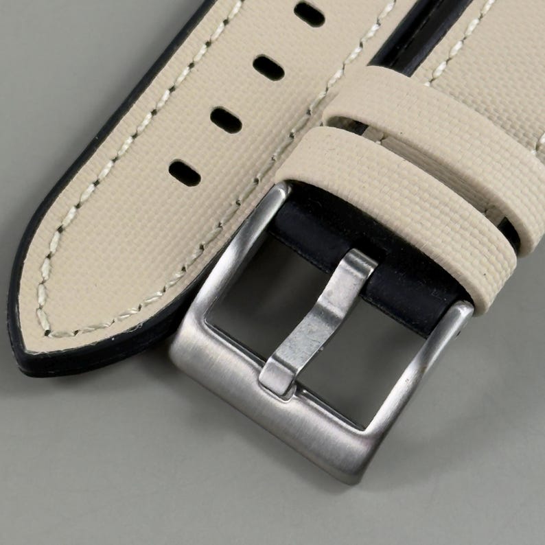 May include: Close-up of a watch strap with a cream-colored canvas exterior and black leather backing. The strap features a silver-colored buckle and contrasting white stitching. The strap has several holes for adjustment.