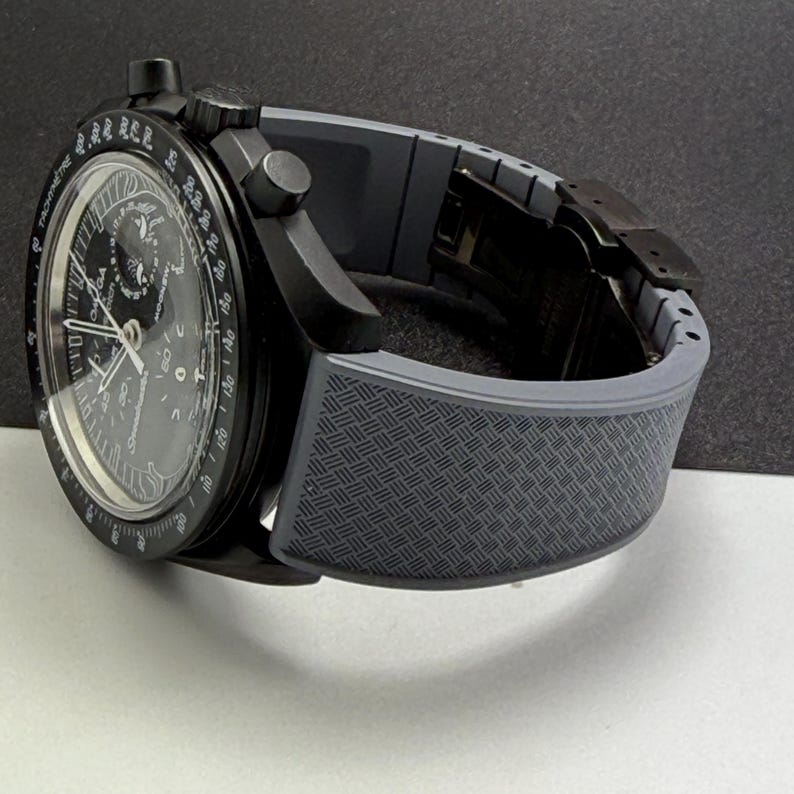 May include: A black wristwatch with a gray patterned band. The watch face is black with white and silver details, including multiple dials and hands. The watch has a sleek, modern design.