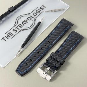 Puede incluir: Una correa de reloj negra con superficie texturizada y costuras azules, con una hebilla de metal plateado. Se incluyen una herramienta para pasadores negra y dos pasadores met&aacute;licos, junto a un estuche transparente con THE STRAPOLOGIST.