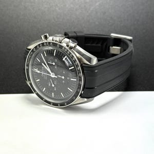 Puede incluir: Un reloj cron&oacute;grafo Omega Speedmaster de acero inoxidable con esfera negra y correa de caucho negra. El reloj presenta m&uacute;ltiples subesferas y un bisel taquim&eacute;trico. El reloj se muestra sobre una superficie blanca y negra.