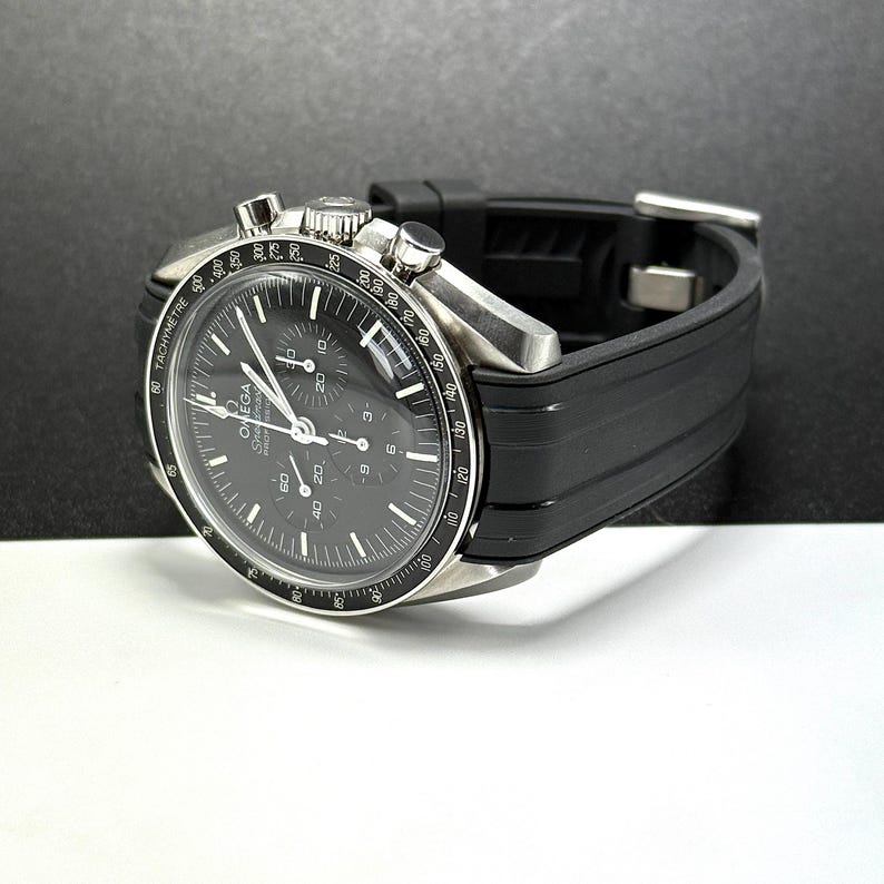 Puede incluir: Un reloj cron&oacute;grafo Omega Speedmaster de acero inoxidable con esfera negra y correa de caucho negra. El reloj presenta m&uacute;ltiples subesferas y un bisel taquim&eacute;trico. El reloj se muestra sobre una superficie blanca y negra.