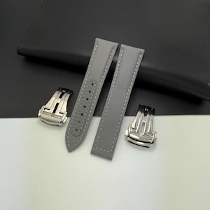 May include: A pair of grey watch straps with white stitching and two silver clasps. The straps are made of a smooth material and have adjustment holes. The clasps are a shiny silver colour.