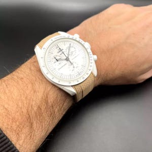 以下が含まれることがあります： ベージュのストラップが付いた白いクロノグラフ腕時計。時計の文字盤にはタキメータースケールと「Speedmaster Professional」の文字が印刷されています。