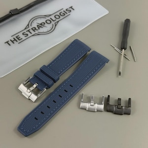 May include: A navy blue watch strap with a textured surface, a silver buckle, and a matching strap. The image also includes a small screwdriver, a pouch with the text "THE STRAPOLOGIST", and two additional clasps.