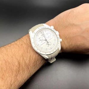 May include: A white wristwatch with a white strap and a white face. The watch features a tachymeter scale around the edge of the face and a chronograph function. The watch is worn on a person's wrist.