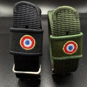 Puede incluir: Dos correas de reloj tejidas, una negra y otra verde oliva, cada una con una trabilla de tela rectangular bordada con un emblema circular de escarapela francesa en rojo, blanco y azul. Las correas incluyen hebillas de metal plateado.
