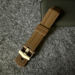 Puede incluir: Una correa de reloj marr&oacute;n con una hebilla plateada. La correa est&aacute; hecha de un material flexible y acanalado. La hebilla tiene forma rectangular con cierre de pasador. La correa del reloj est&aacute; sobre una superficie gris.