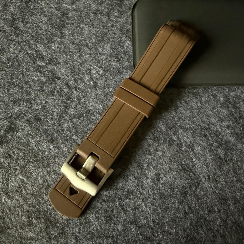 Puede incluir: Una correa de reloj marr&oacute;n con una hebilla plateada. La correa est&aacute; hecha de un material flexible y acanalado. La hebilla tiene forma rectangular con cierre de pasador. La correa del reloj est&aacute; sobre una superficie gris.