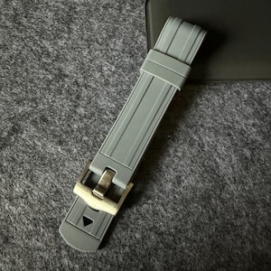 Puede incluir: Una correa de reloj gris con una hebilla plateada. La correa est&aacute; hecha de un material flexible y presenta un dise&ntilde;o texturizado con l&iacute;neas paralelas. La hebilla tiene forma rectangular y cierre de pasador.