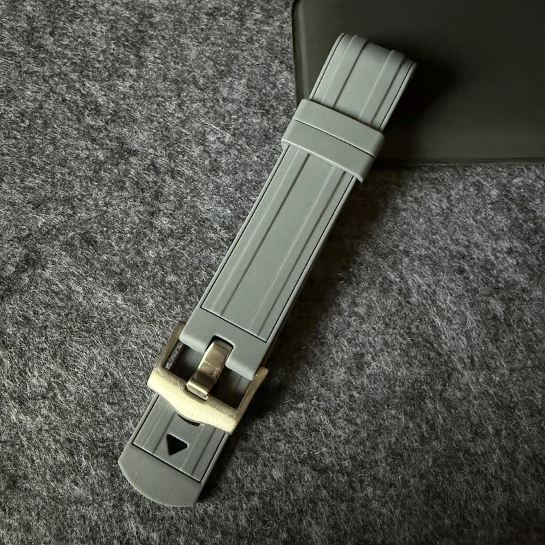 Puede incluir: Una correa de reloj gris con una hebilla plateada. La correa est&aacute; hecha de un material flexible y presenta un dise&ntilde;o texturizado con l&iacute;neas paralelas. La hebilla tiene forma rectangular y cierre de pasador.